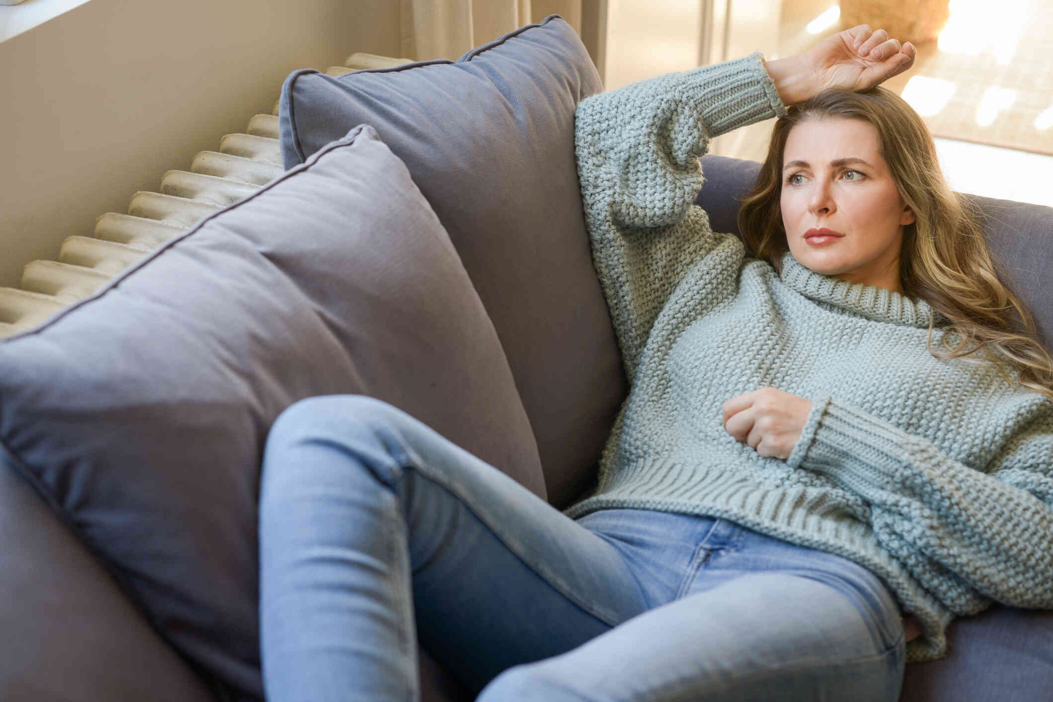 A woman in a green sweater lays on her back on a grey couch and gazes off with a sad expression.